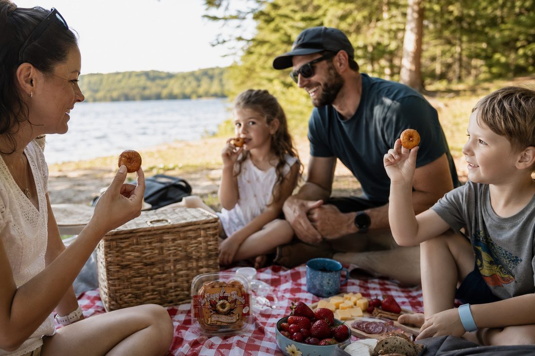 Famille souriante qui savoure des beignets Le Temps d'une Bouchée lors d'un pique-nique au bord d'un lac